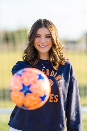 A person wearing a navy blue sweatshirt with orange lettering poses with a blurred football in the foreground.