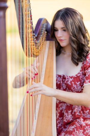 A harpist in a red floral dress performs elegantly on a classical pedal harp in an outdoor setting.