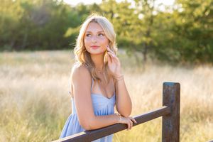 Blonde woman in light blue dress leaning on rustic wooden fence in golden field during sunset.