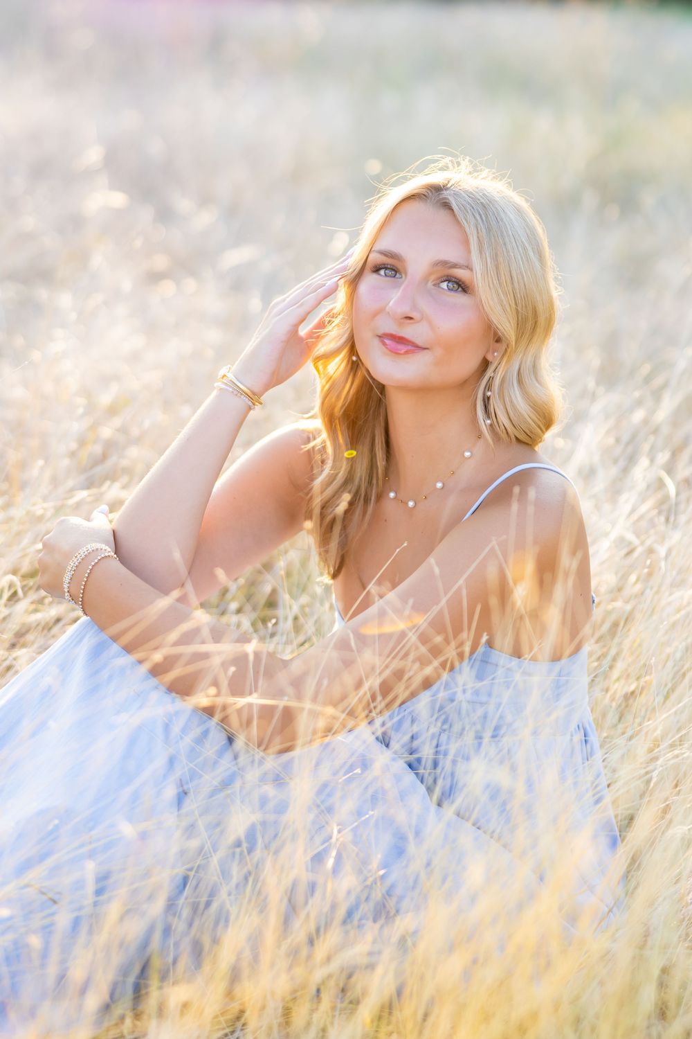 Young blonde woman in a light blue dress sitting in golden wheat field, bathed in warm sunlight.