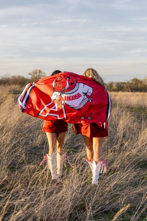 People wearing Arkansas sports gear bend down playfully in tall grass field.