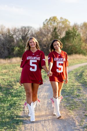 Two people in Arkansas sports jerseys walk together on a dirt path at sunset.