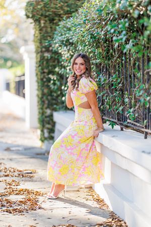A person in a yellow and pink floral dress twirls playfully on a sidewalk with fallen autumn leaves and greenery.