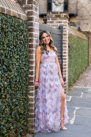 A person wearing a flowing lavender floral maxi dress poses against a brick wall with ivy in an outdoor setting.