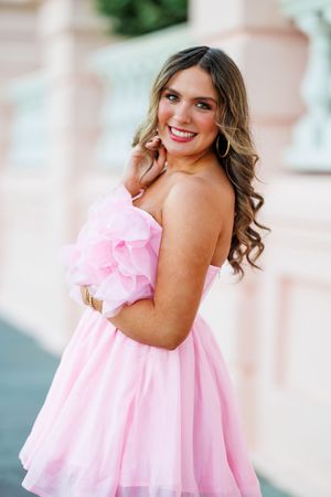 A pink strapless mini dress with ruffled details and a flared skirt photographed against a blurred outdoor background.