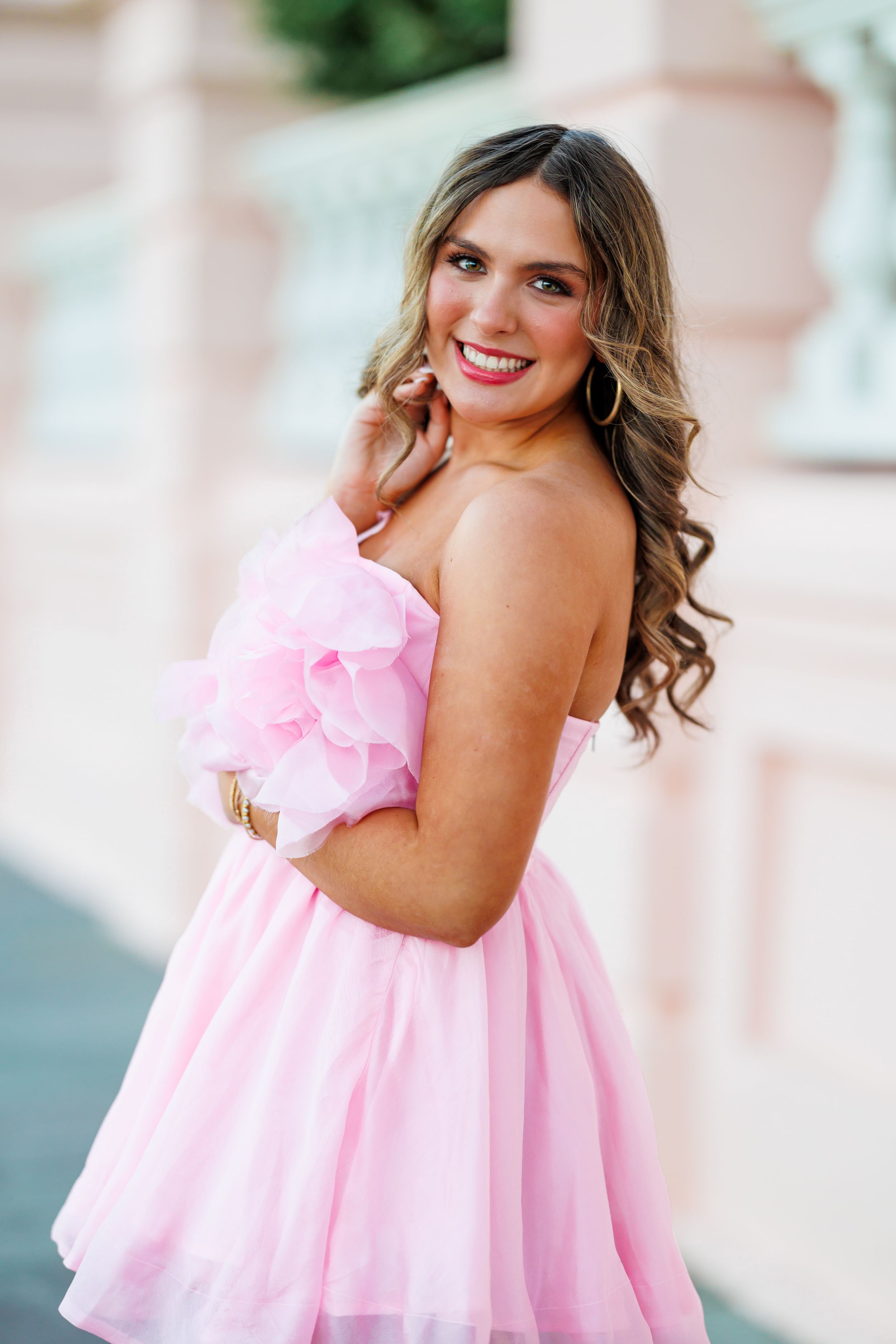 A pink strapless mini dress with ruffled details and a flared skirt photographed against a blurred outdoor background.