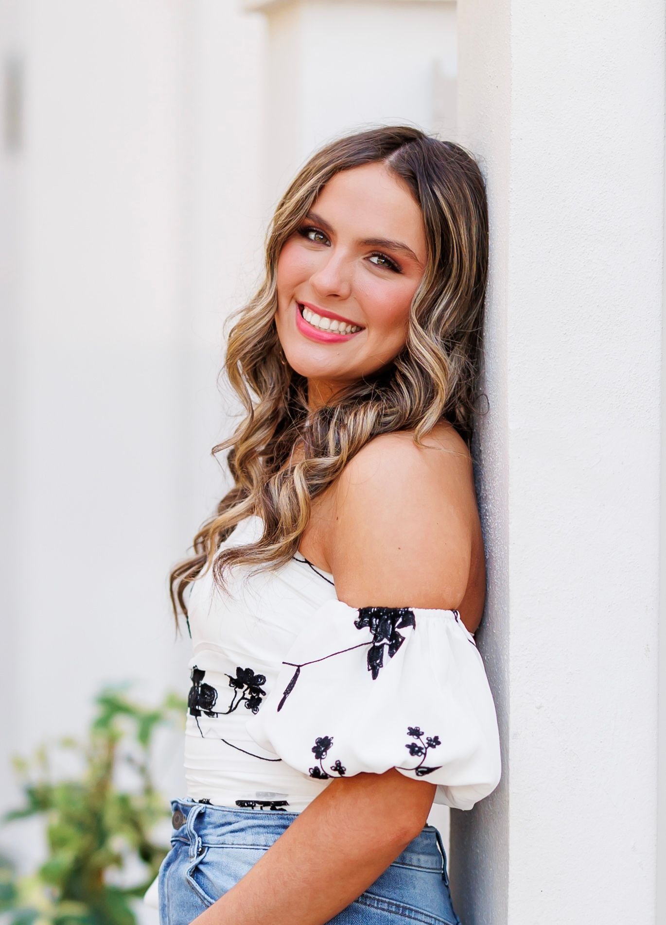 Woman in white off-shoulder top and denim smiles while posing against light colored wall with greenery.