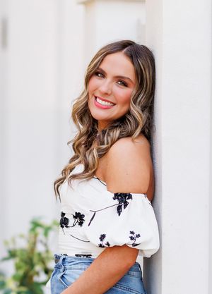 Woman in white off-shoulder top and denim smiles while posing against light colored wall with greenery.