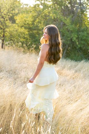 Young woman in white ruffled dress standing in golden field with long wavy hair, bathed in warm sunlight.