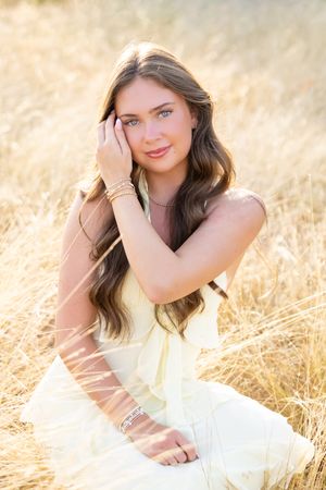 Young woman in yellow dress sitting in golden wheat field, wearing bracelets, with long brown hair and blue eyes.