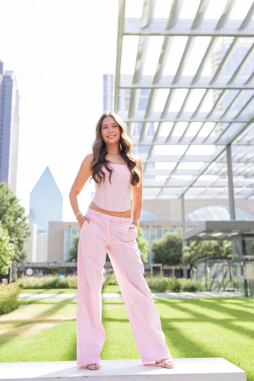 Young woman in pink crop top and wide-leg pants poses confidently outdoors near modern building with city skyline.