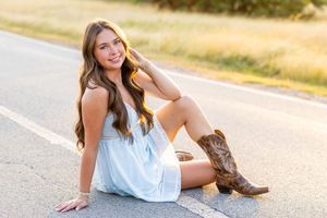 A smiling young woman in a white dress and cowboy boots sits on a road, posing for senior portraits.