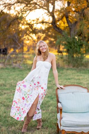 Model in floral maxi dress poses next to vintage chair in outdoor garden setting.