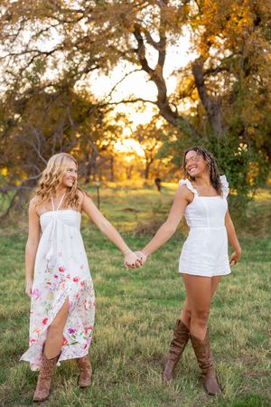 Two friends in white dresses and boots hold hands in sunset garden setting.