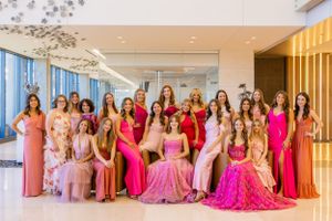 Large group photo of people wearing formal pink and red gowns in an elegant hallway.