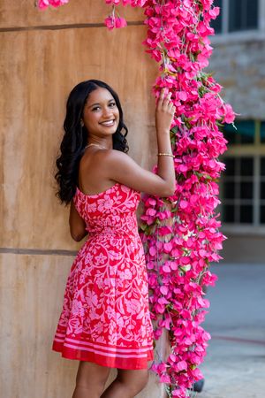 A woman in a red floral dress poses beside vibrant pink bougainvillea flowers in an outdoor setting.