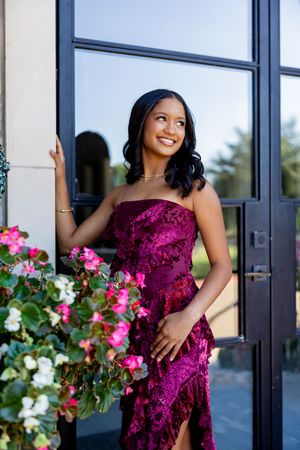 Woman in elegant burgundy velvet gown poses by doorway with pink flowers in outdoor setting.
