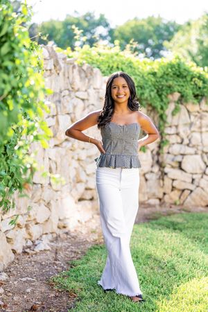 A woman in a black and white outfit poses gracefully against a stone wall in an outdoor garden setting.