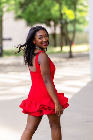 Young Black woman in red ruffled mini dress smiling outdoors, hair flowing, wearing beaded necklace on sunny day.