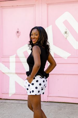 Smiling woman in black top and polka dot shorts posing in front of a pink wall.