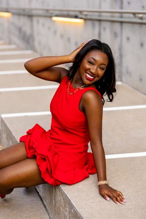 A young Black woman in a red dress sits on steps, smiling brightly, wearing gold jewelry and accessories.