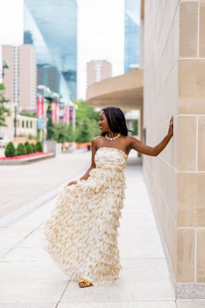 A smiling woman in a gold ruffled strapless gown poses against a stone wall in an urban setting.