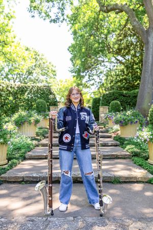 A person wearing a varsity jacket and jeans stands on a garden path surrounded by greenery and stone posts.