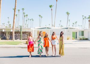 Women holding hands while walking down palm tree-lined street in bright dresses.