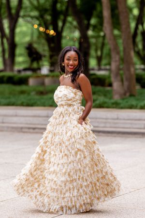 A smiling woman twirls in a stunning gold and white ruffled strapless ball gown in an outdoor garden setting.