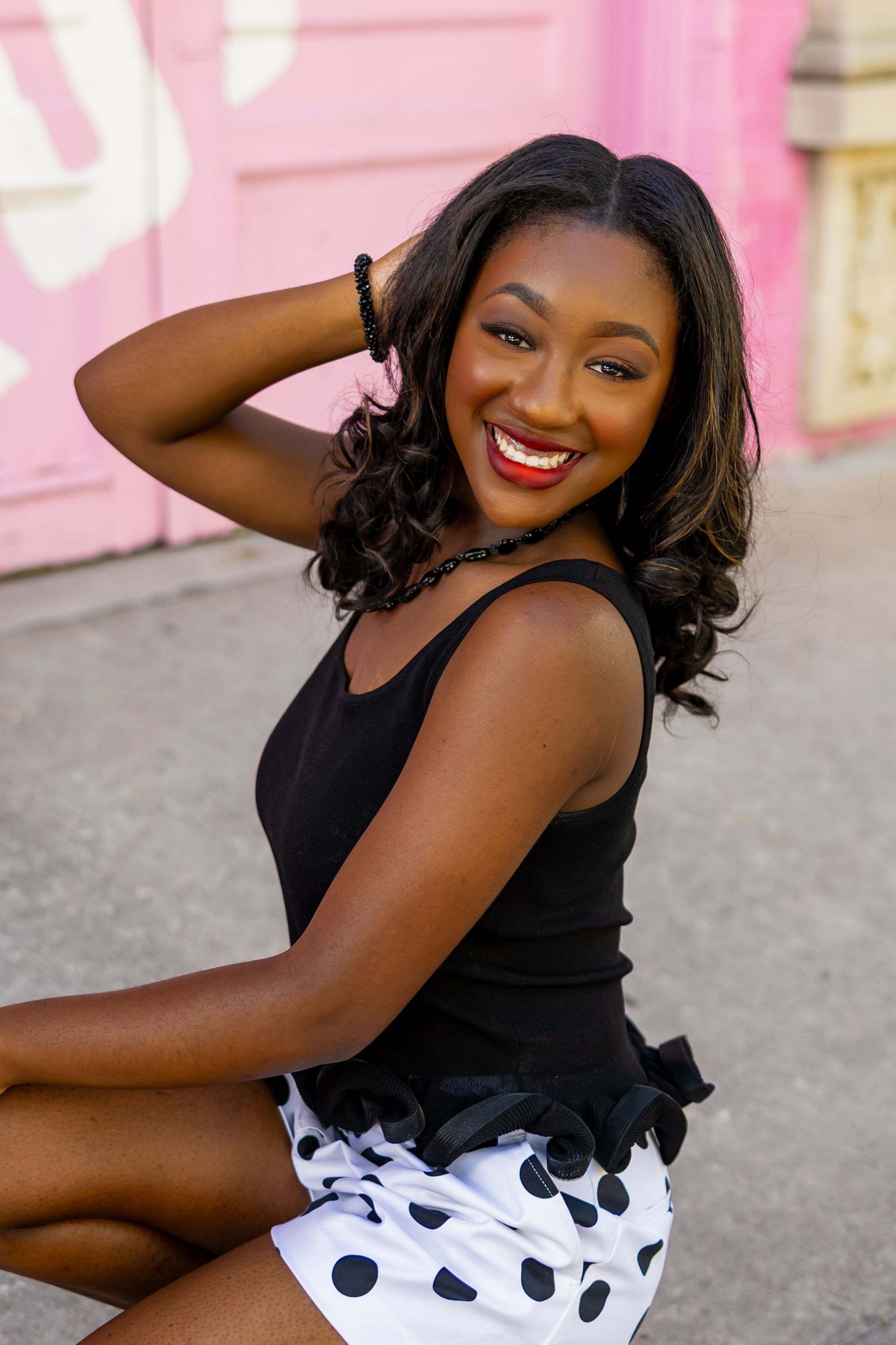 Smiling young Black woman in black top and polka dot skirt sitting outdoors against a pink wall.