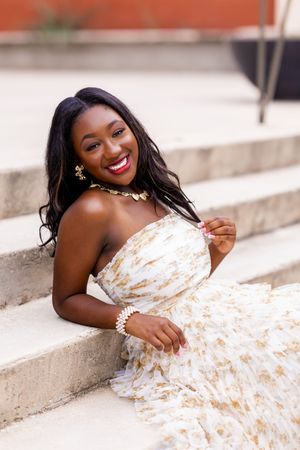 Young Black woman in white and gold strapless gown sitting on stone steps, smiling, wearing gold jewelry.