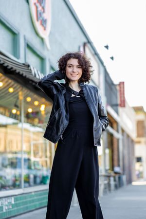Person in black coat strikes playful poses against urban storefront backdrop.