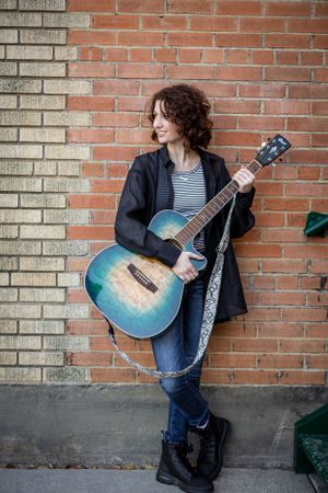 Person with curly hair plays blue acoustic guitar against brick wall background.