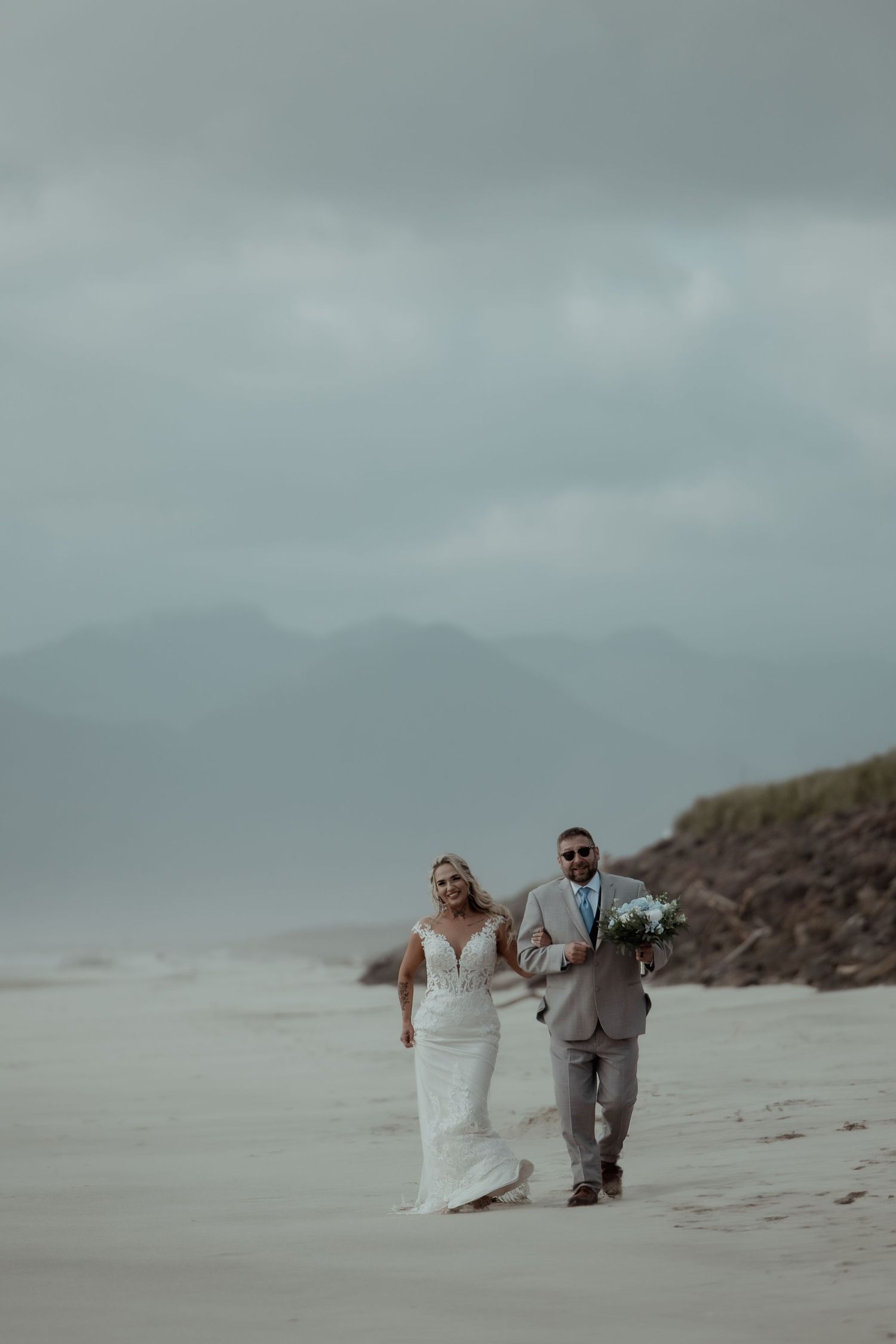 A couple walking together on a misty beach with mountains in the background on their wedding day.