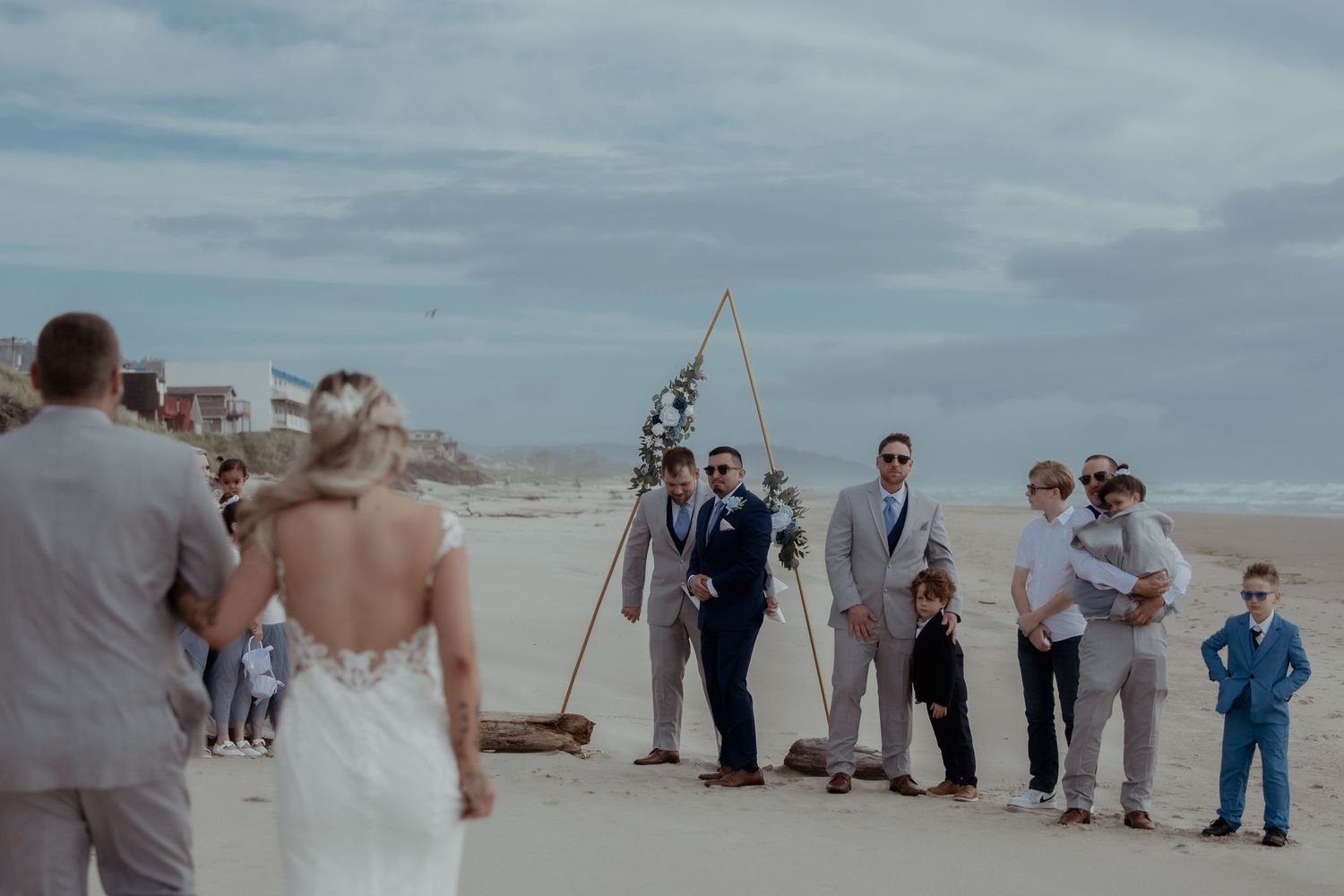 A beachside wedding ceremony setup with guests gathering along the shoreline as waves roll in on a cloudy day.