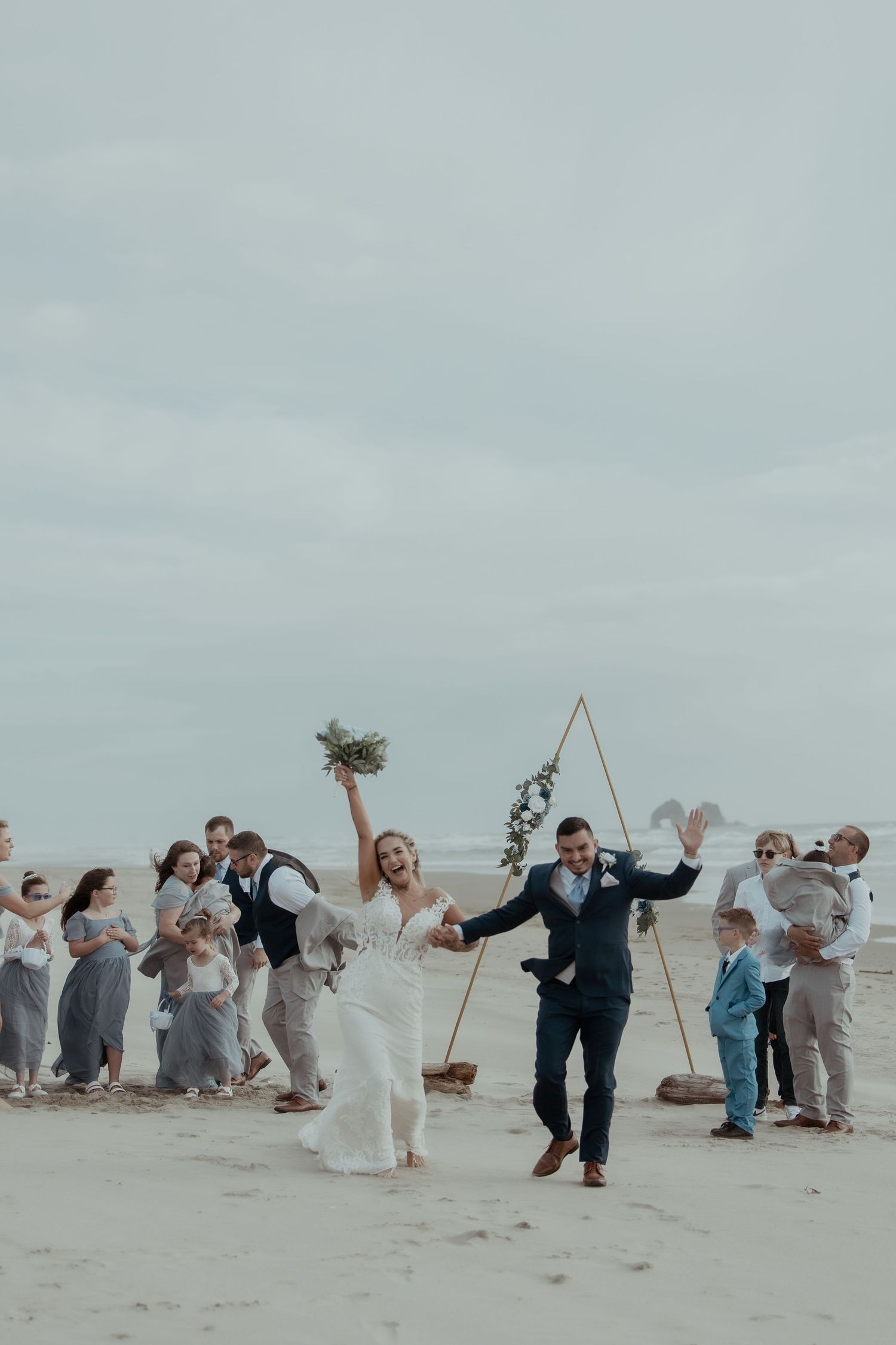 A wedding party enjoys playful festivities on a misty beach with large decorative geometric shapes.