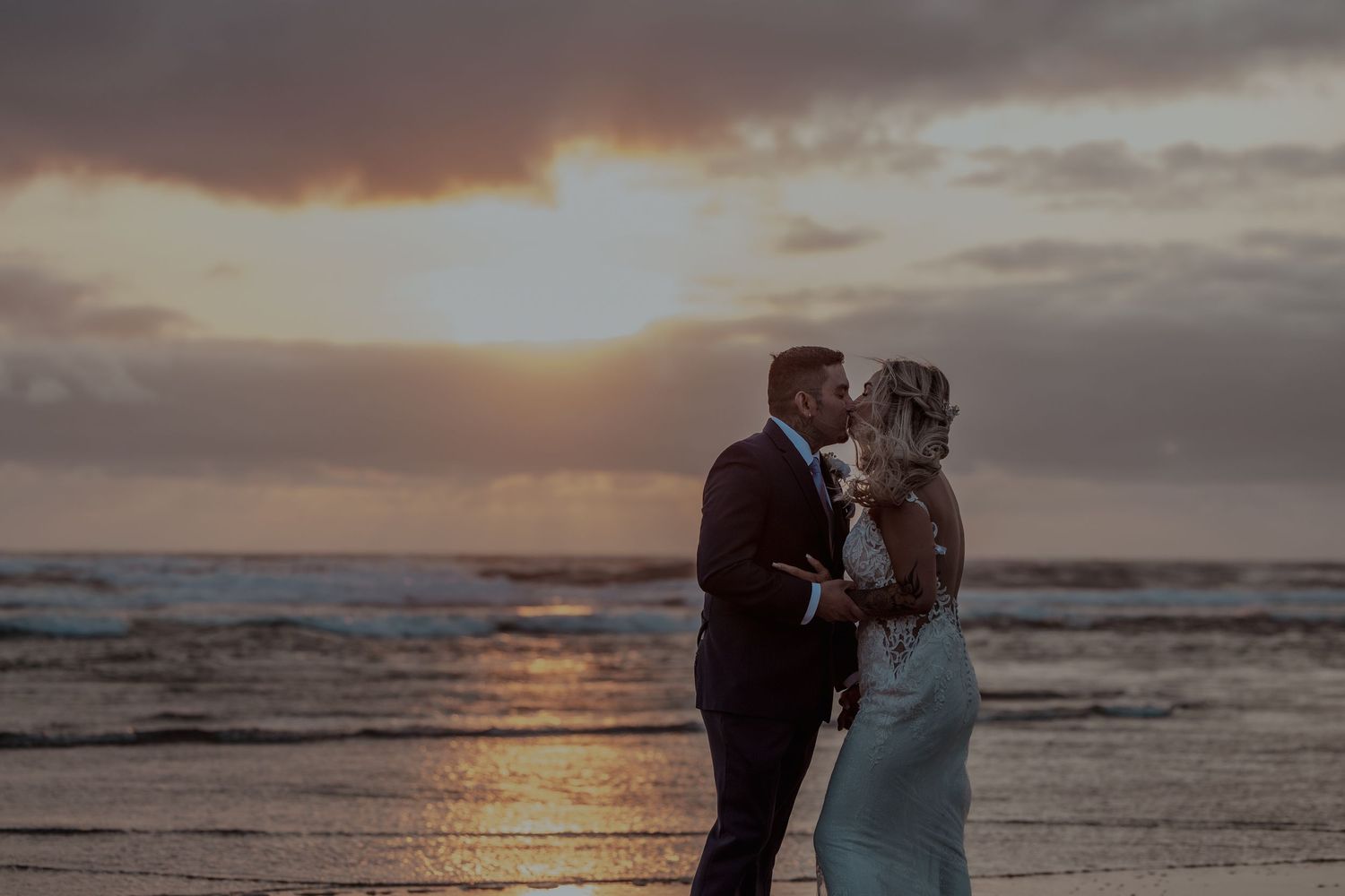 A romantic couple embraces on the beach at sunset with dramatic orange clouds reflecting on the ocean waves.