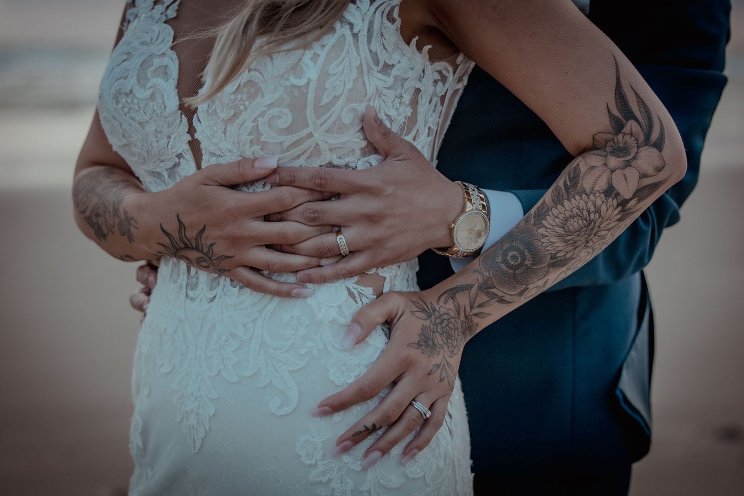 Close-up of tattooed arms embracing a person wearing a white lace wedding dress during a romantic moment.