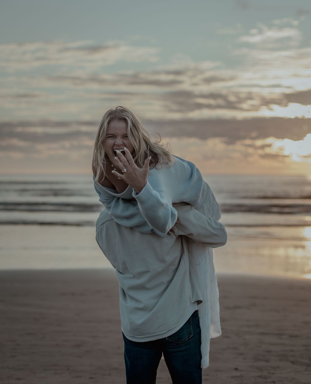 A person in an oversized sweater laughs joyfully on a beach at sunset with dramatic cloudy skies in the background.