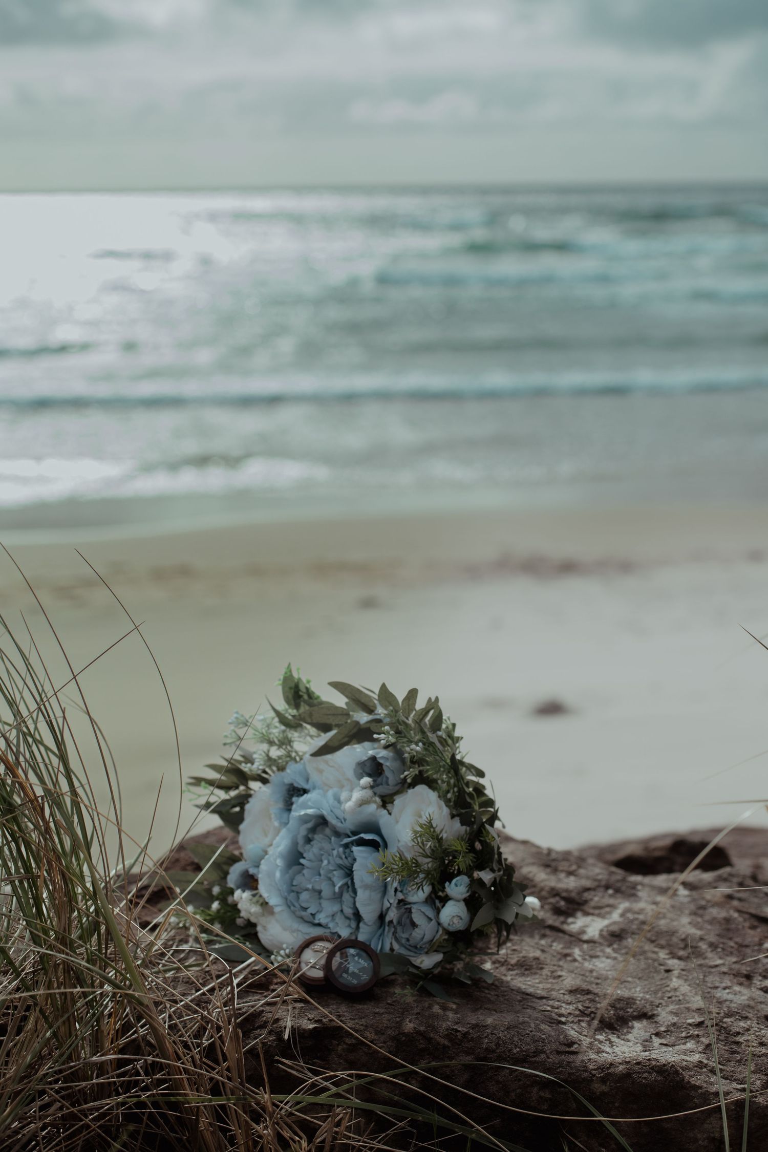 A soft blue bridal bouquet rests on driftwood at the beach with ocean waves and moody sky in the background.