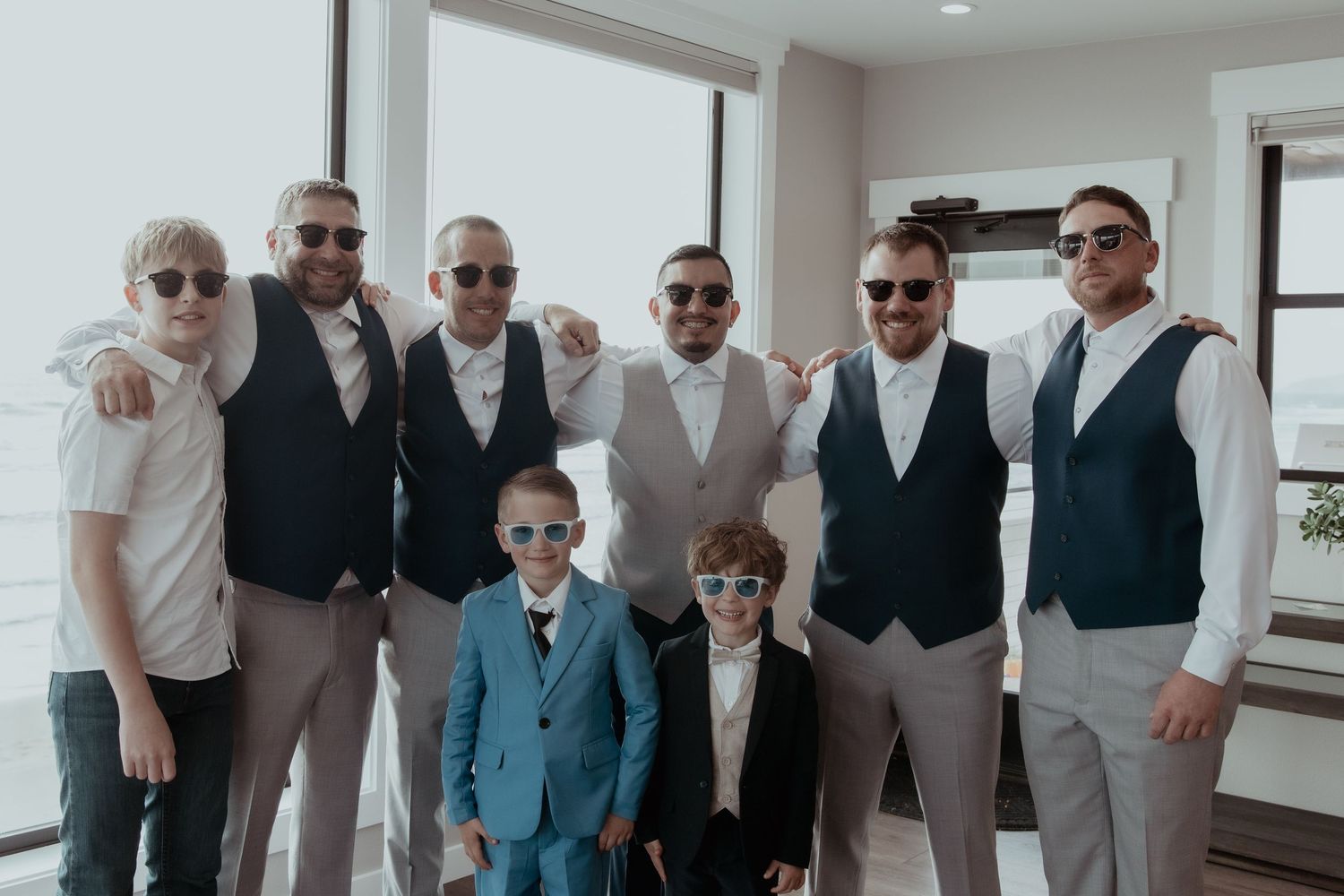 A group in formal attire wearing sunglasses pose together wearing vests and dress clothes in a bright interior space.