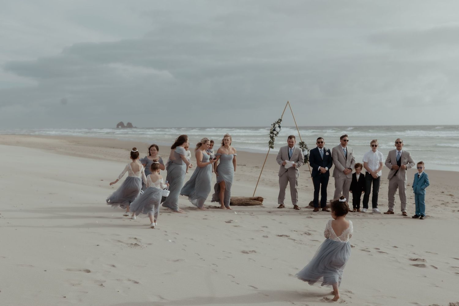 A wedding party in soft blue and gray attire poses on a misty beach with cloudy skies overhead.