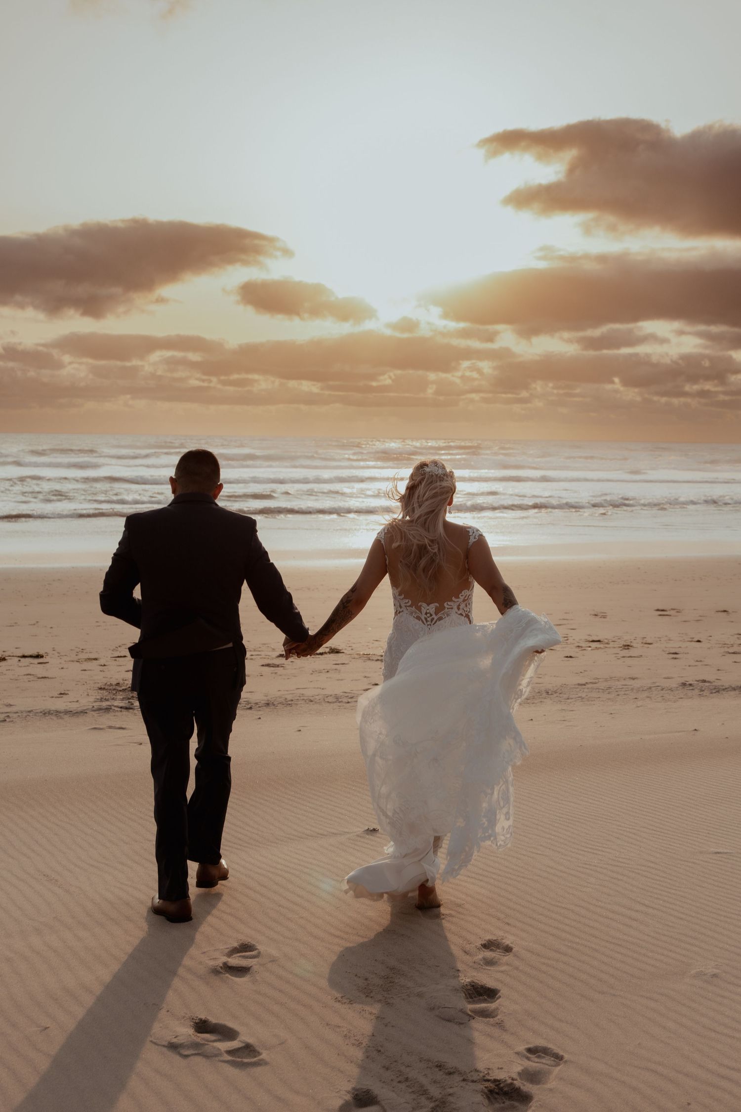 A newlywed couple walks hand in hand on a beach during golden sunset hour, leaving footprints in the sand.