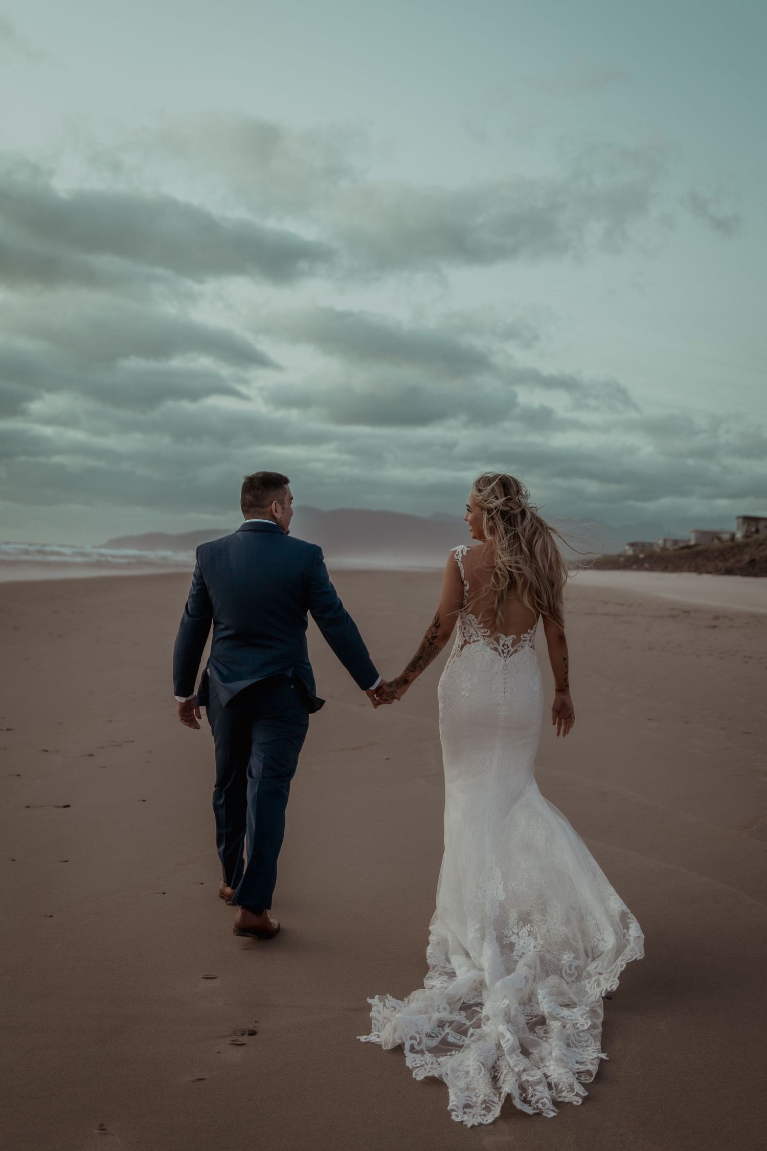 Romantic newlyweds walking together on a moody beach at sunset with cloudy skies and footprints in the sand.