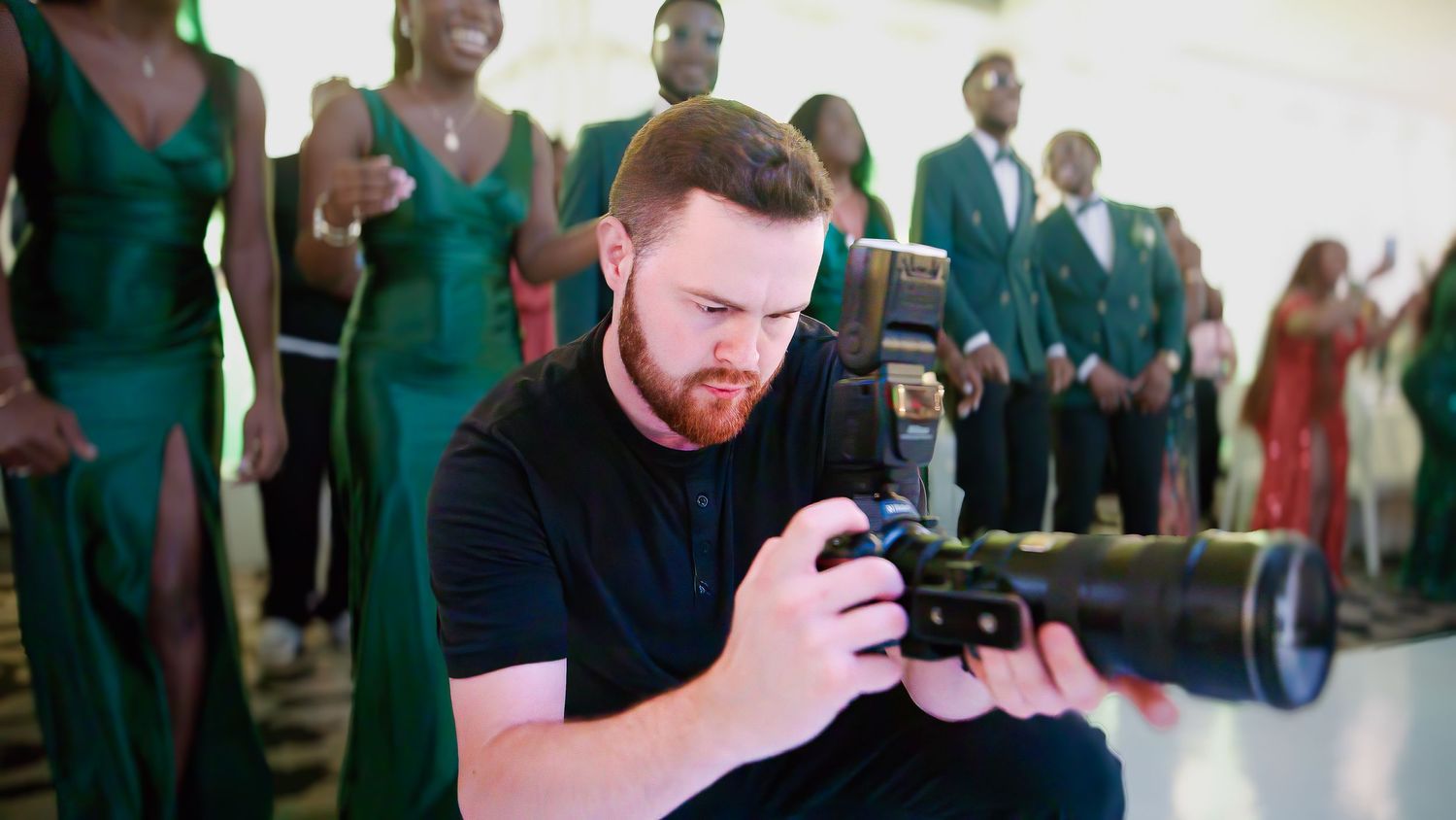 Professional photographer with red beard in black t-shirt uses camera with telephoto lens at formal event with green-clad group.