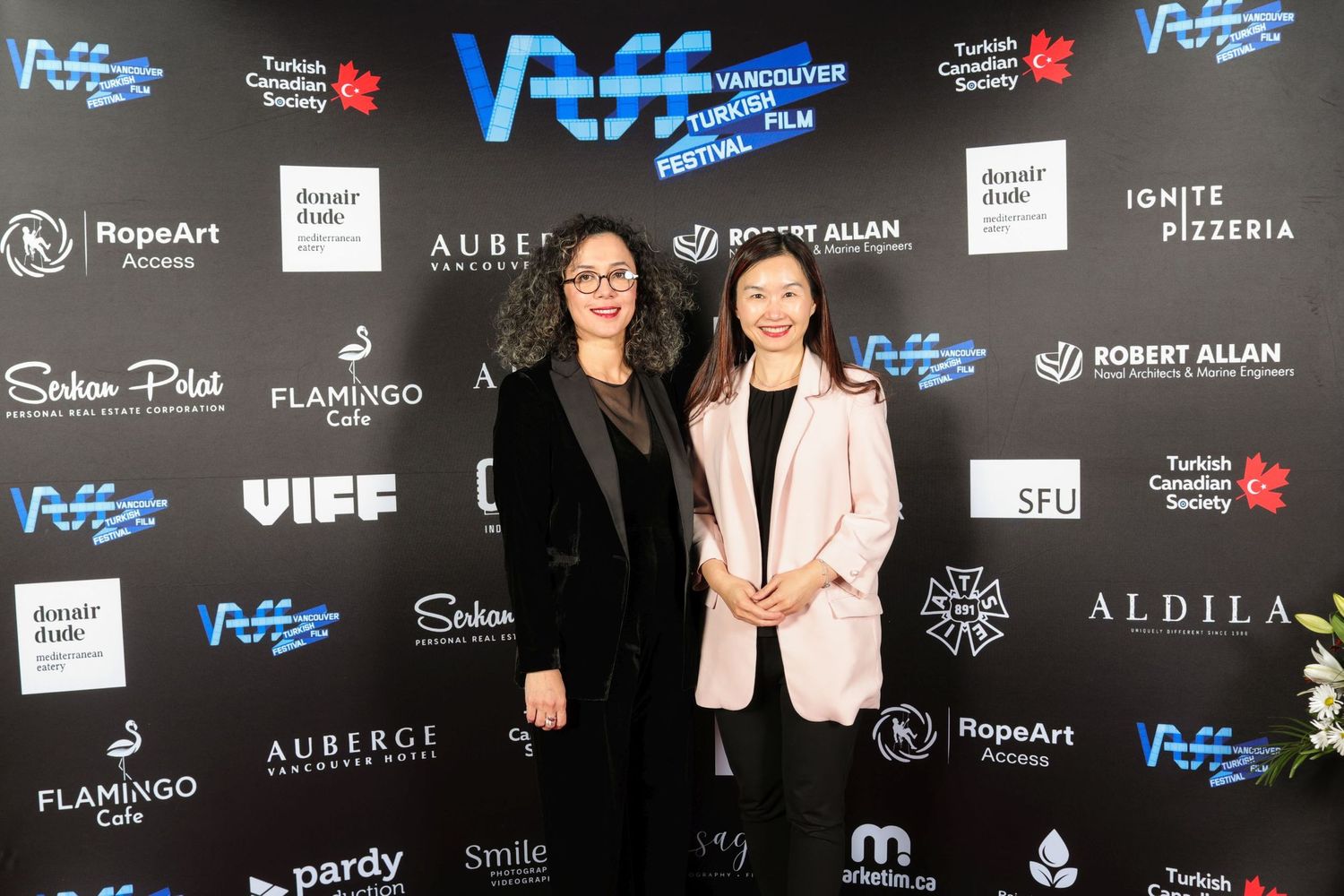 Two people pose together at a media event with blue logos and sponsor banners visible in the background.