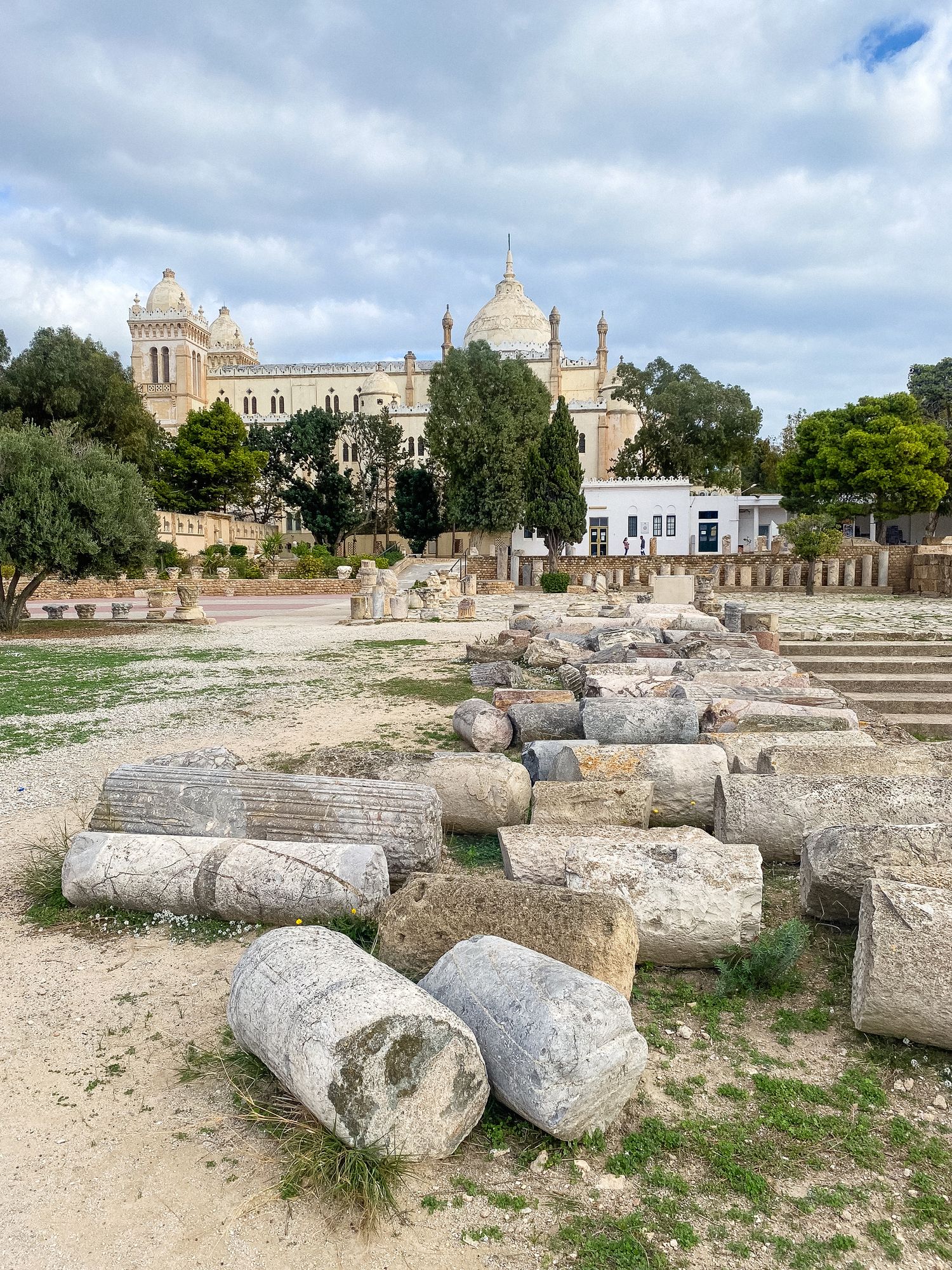 Ancient stone columns and ruins in foreground with ornate church building and blue sky in background at archaeological site.