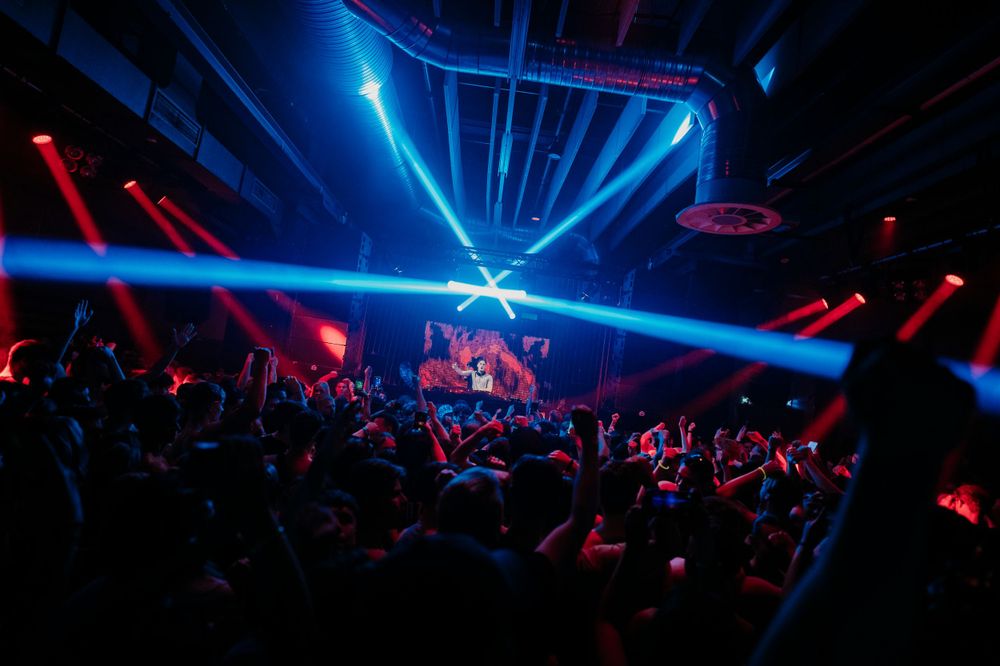 Blue laser lights sweep across a dark concert venue filled with silhouetted crowd.