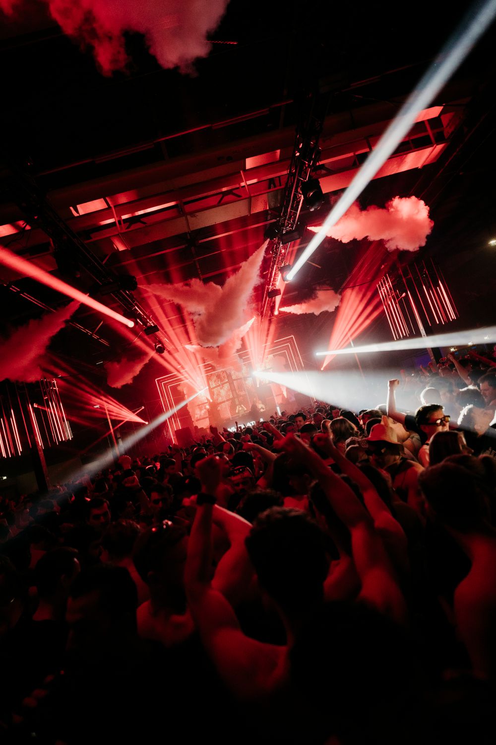 Spectacular light show with white beams cutting through red smoke at a concert venue.