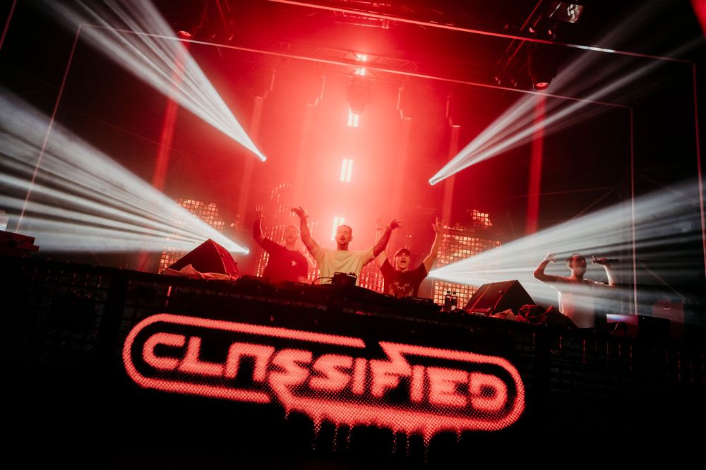 Concert venue illuminated by dramatic white spotlights and red ambient lighting during electronic music show.